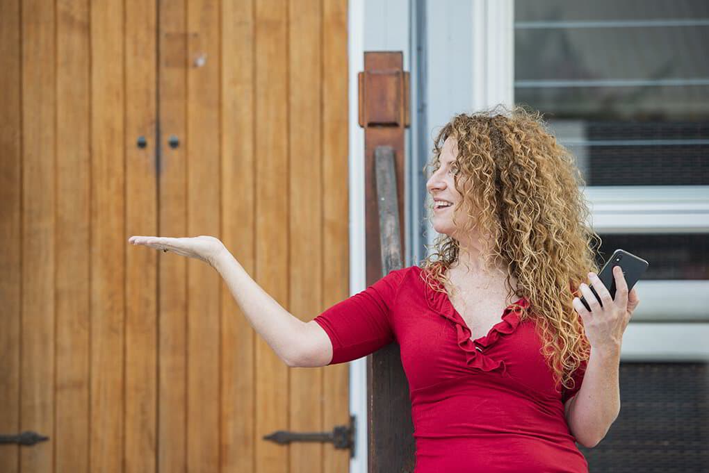 Lisa Monks smiling and gesturing with an open palm, phone in hand, on a timber verandah