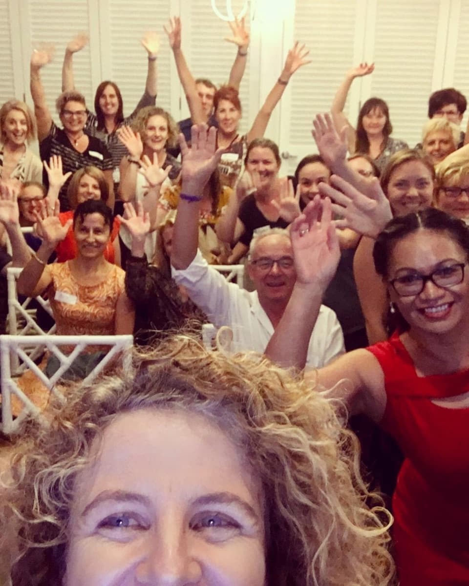 A full workshop room of smiling small business owners with their hands in the air at one of Lisa's training sessions