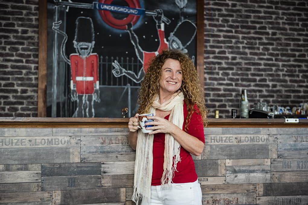 Lisa Monks holding a coffee mug and smiling at a rustic café bar